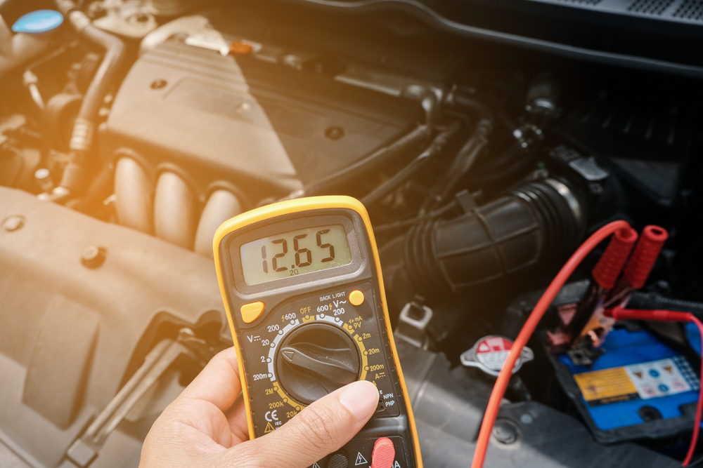 A person using a yellow multimeter with red test lead to check a car's electrical system.